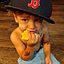 child, boy, baseball_cap, hat, cheese, eating, shirtless, shorts, floor, wooden_floor, tattoo, hand, curly_hair, indoor, playful, smiling, cute, young_child, portrait, casual
