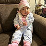 baby, child, infant, pink_socks, tracksuit, hat, armchair, brown_furniture, indoor, curious_expression, face, hands, clothing, laundry_basket, cozy, home, seated, portrait, casual, young_child