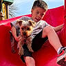 boy, child, dog, small_dog, slide, playground, outdoor, red_slide, casual_clothing, shorts, sneakers, daylight, fun, pet, animal, play, happy, cute, summer, leisure