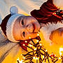 baby, child, smiling, face, santa_hat, christmas_lights, holiday, festive, blanket, soft, cute, happy, warm_light, indoors, cozy, red_clothing, white_fur, portrait, close_up, celebration