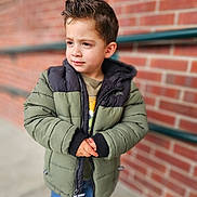 Pierre is registered to the contest to win money with this photo: child, boy, jacket, green_jacket, black_jacket, hair, curly_hair, standing, outdoor, brick_wall, railing, casual_clothing, jeans, portrait, expression, young, hands, pensive, urban, cold_weather