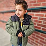 child, boy, jacket, green_jacket, black_jacket, hair, curly_hair, standing, outdoor, brick_wall, railing, casual_clothing, jeans, portrait, expression, young, hands, pensive, urban, cold_weather