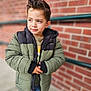 Pierre is registered to the contest to win money with this photo: child, boy, jacket, green_jacket, black_jacket, spiky_hair, brick_wall, outdoor, casual_clothing, portrait, winter_clothing, young_child, standing, looking_away, curious_expression, hands_together, blue_jeans, urban, daylight, cool_weather