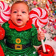 Elio joined the competition — help win amazing prizes! baby, child, elf_costume, green_clothing, red_collar, peppermint_lollipop, holiday_decor, christmas, festive, snow, red_ornament, face, smile, happy, celebration, cute, portrait, indoor, colorful, sparkly
