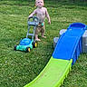 toddler, child, grass, outdoor, toy, slide, lawn_mower, barefoot, pacifier, necklace, summer, fence, backyard, playtime, greenery, shorts, sunlight, young_child, playground, garden