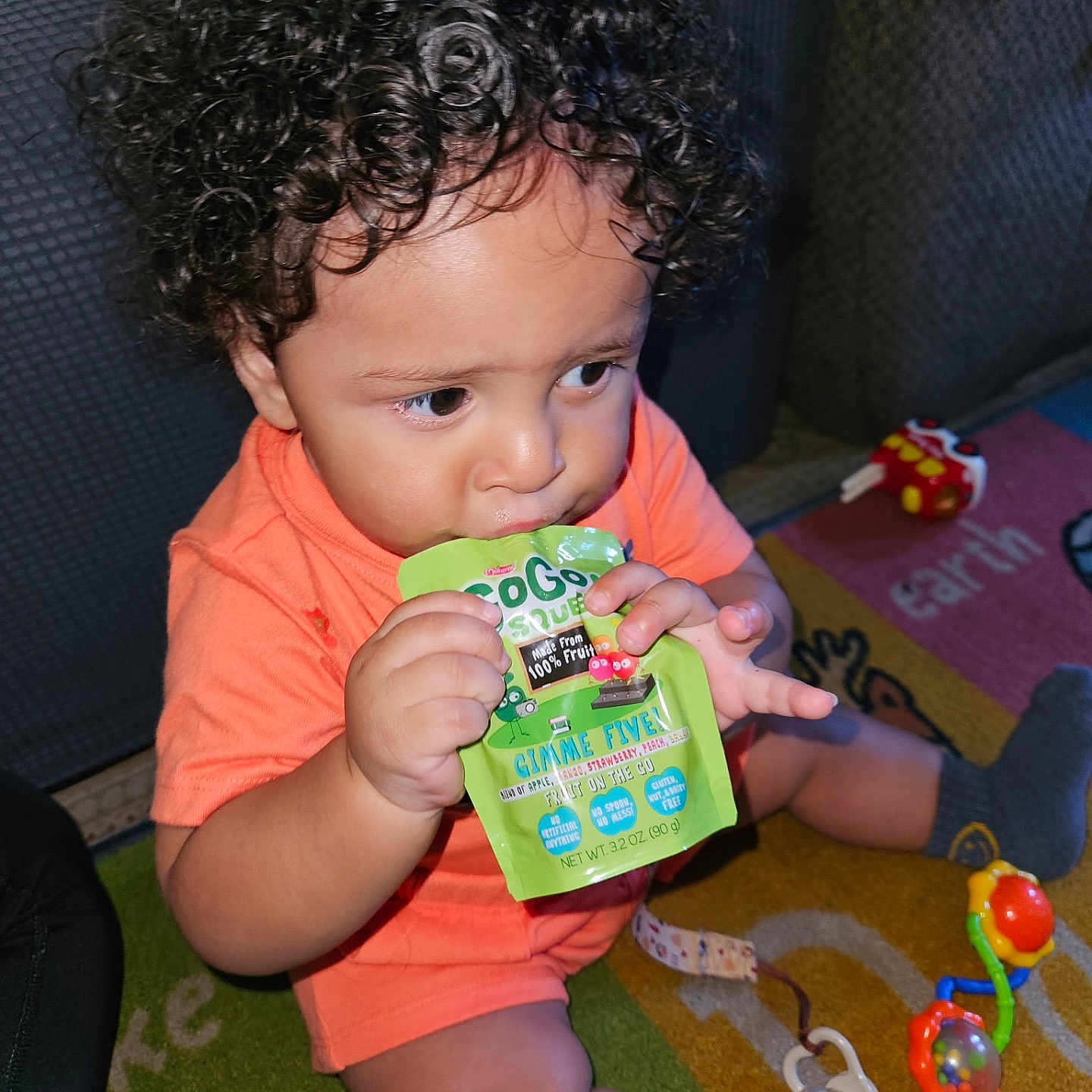 Khyaire is registered to the contest to win money with this photo: baby, candid, carpet, child, colorful, curly_hair, cute, floor, focus, fruit_pouch, hand, indoor, orange_shirt, pacifier, playing, sitting, snack_time, socks, toddler, toy