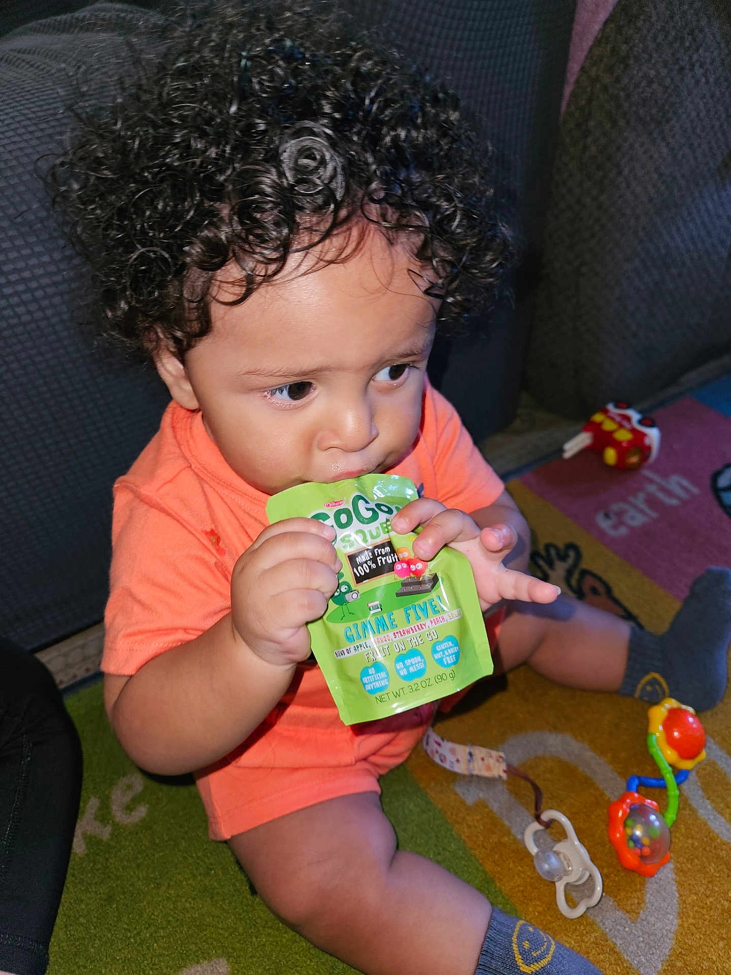 Khyaire is registered to the contest to win money with this photo: toddler, child, curly_hair, orange_shirt, fruit_pouch, sitting, carpet, toy, pacifier, indoor, snack_time, cute, baby, playing, floor, colorful, socks, focus, hand, candid