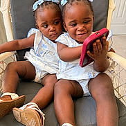Delilah And Dahlia is registered to the contest to win money with this photo: toddler, child, phone, chair, blue_bow, matching_outfits, white_sandals, indoor, cushion, smile, curious, two_children, baby_clothing, braided_hair, playful, cute, happy, person, home, technology
