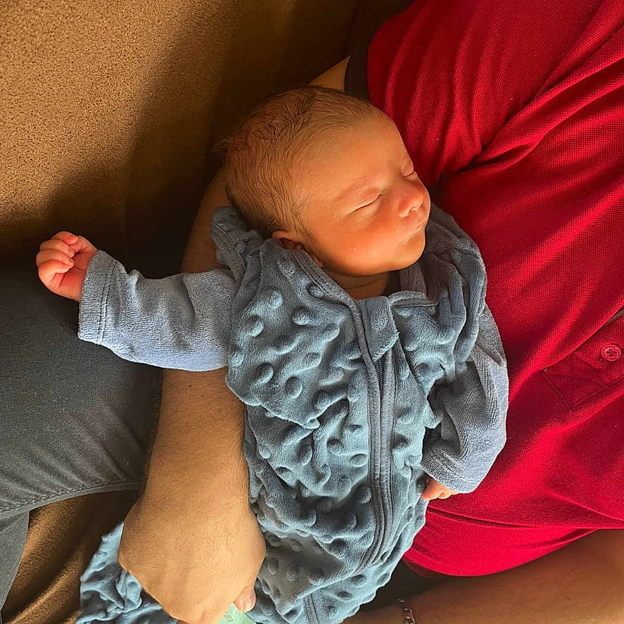 Raphaël a rejoint le concours — aidez-le/la à gagner de superbes lots ! adult_arm, baby, blue_onesie, caring, closeup, comfort, cozy, hand, indoor, infant, newborn, pacifier, peaceful, person, portrait, red_shirt, rest, sleeping, tender, texture