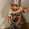 baby, child, toddler, portrait, standing, smiling, indoors, hat, chef_hat, shirt, graphic_shirt, pants, shoes, adult_hand, hand_holding, wooden_floor, wall, home, fashion, candid