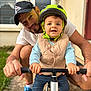 Klarence a rejoint le concours — aidez-le/la à gagner de superbes lots ! adult, bike, blue_shoes, building, cap, casual_clothing, child, face, grass, hand, helmet, outdoor, pavement, person, safety_gear, smiling, support, toddler, vest, window