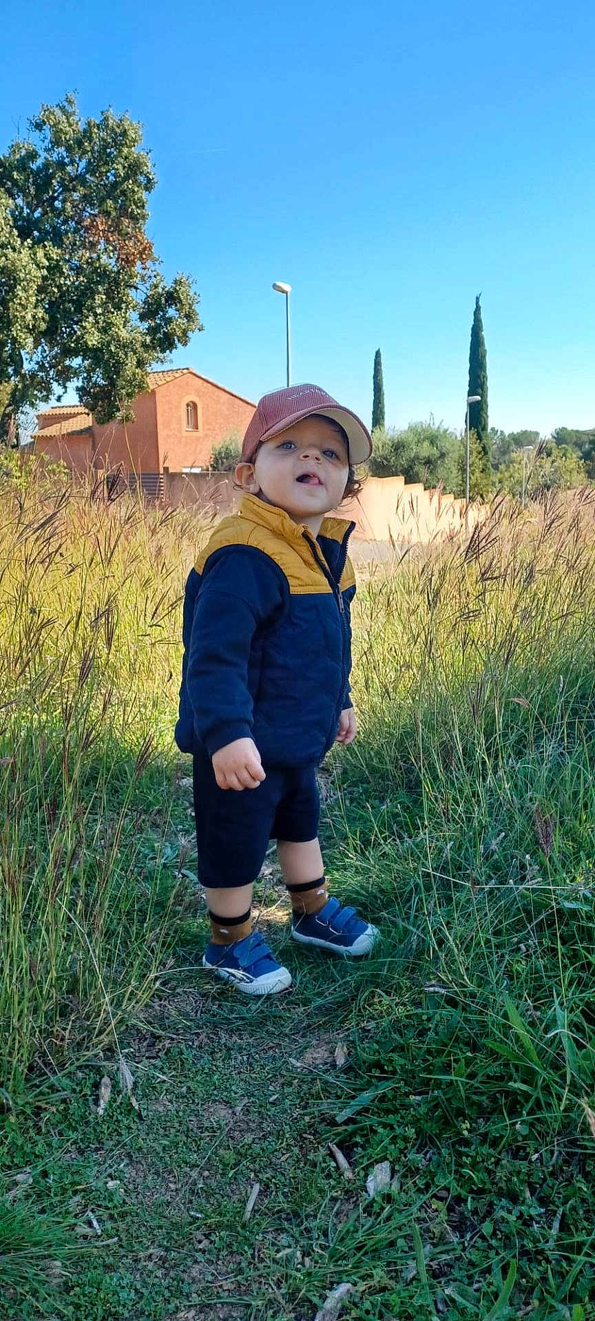 Klarence a rejoint le concours — aidez-le/la à gagner de superbes lots ! blue_sky, cap, child, curious, daylight, grass, jacket, nature, outdoor, path, person, plants, rural, shoes, shorts, smile, standing, sunny, toddler, trees