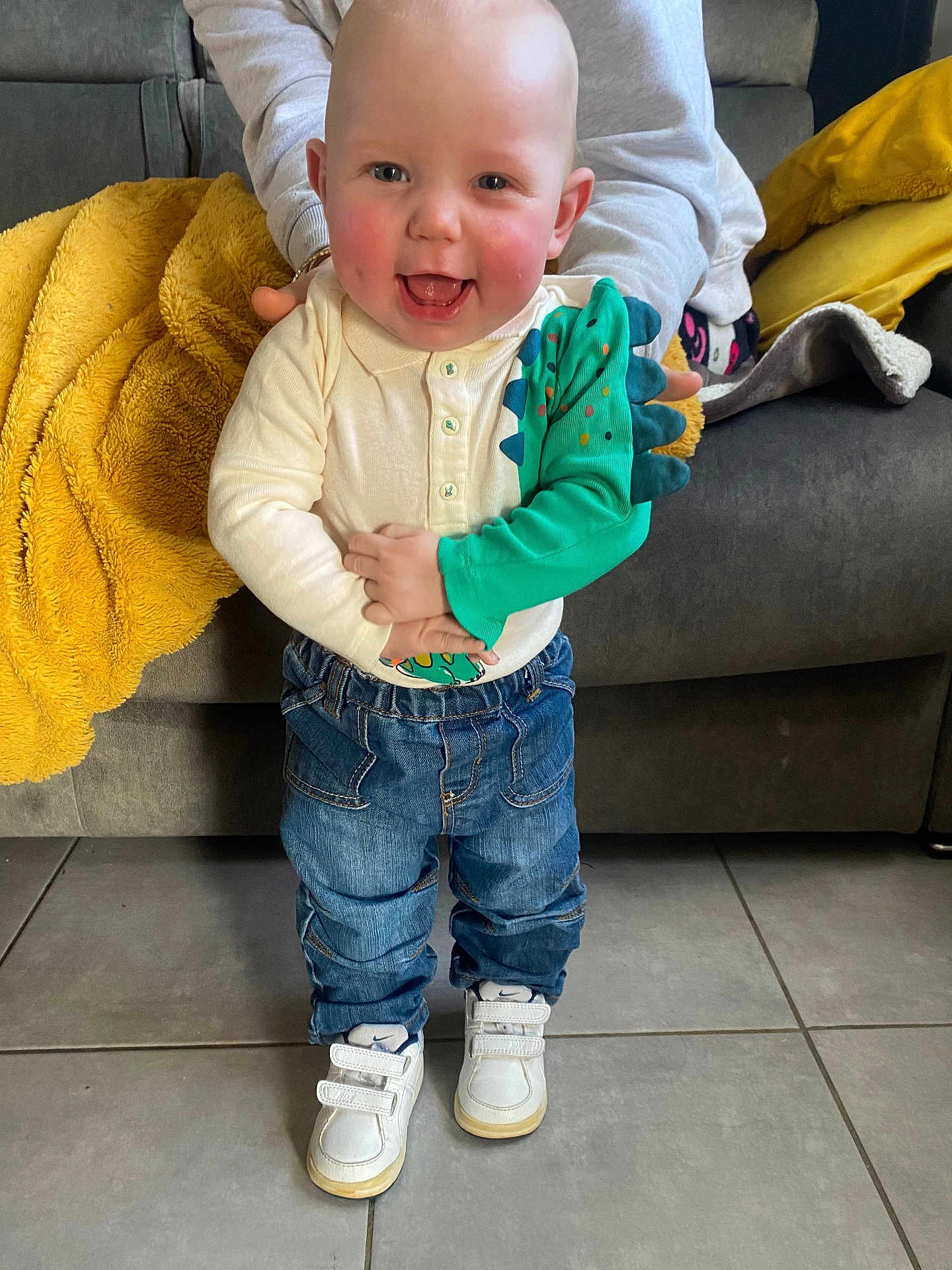 Logan a rejoint le concours — aidez-le/la à gagner de superbes lots ! baby, toddler, smiling, dinosaur_sleeve, jeans, white_sneakers, tile_floor, sofa, mustard_blanket, parent_hand, rosy_cheeks, standing, happy, indoor, casual_clothing, tiny_hands, portrait, living_room, denim, cozy