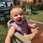 Keira is registered to the contest to win money with this photo: arm, baby, background, carpet, chair, child, dining, expression, foot, happy, high_chair, indoor, light, people, person, purple_clothing, restaurant, smile, table, wooden_table