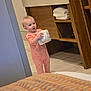 baby, bathroom, cabinetry, child, clothing, curious, floor_tiles, holding, home, indoor, light, person, pink_onesie, standing, tile_floor, toddler, toilet_paper, towels, wooden_cabinet, young_child