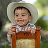 Dylan is registered to the contest to win money with this photo: baby, child, cowboy_hat, smiling, portrait, outdoor, wooden_chair, seated, infant, face, hands, western_clothing, shirt, jeans, nature, tree, greenery, background_blur, happy, photography