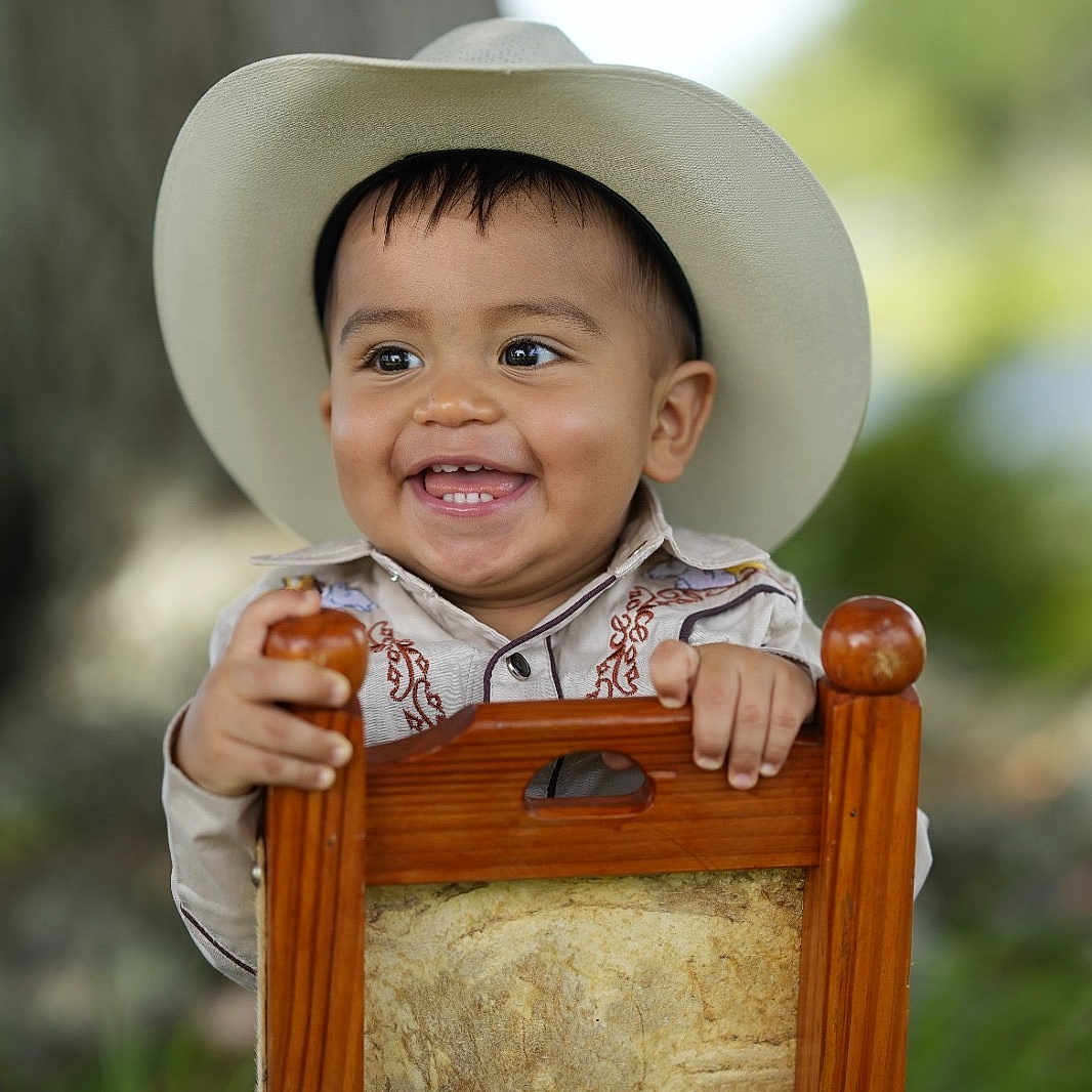 Dylan is registered to the contest to win money with this photo: baby, child, cowboy_hat, smiling, portrait, outdoor, wooden_chair, seated, infant, face, hands, western_clothing, shirt, jeans, nature, tree, greenery, background_blur, happy, photography