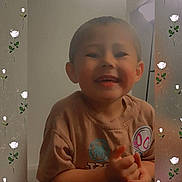 Alakai is registered to the contest to win money with this photo: child, smiling, indoor, portrait, boy, short_hair, casual_clothing, rose_pattern, decorative_frame, happy, cute, young, sitting, wall, room, light, face, hands, tshirt, background