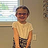 child, girl, smiling, glasses, indoors, window, blinds, patterned_skirt, white_top, carpet, wall, power_outlet, eyeglasses_display, standing, happy, person, hair, earrings, light, room