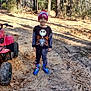 child, young_child, smiling, beanie_hat, sweater, striped_pants, blue_boots, atv, toy_vehicle, dirt_path, sand, forest, pine_trees, sunlight, outdoors, standing, playful, portrait, muddy, leaves