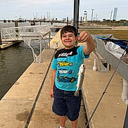 Benjamin is registered to the contest to win money with this photo: child, boy, smile, fish, dock, pier, water, harbor, boats, lighthouse, fishing_rod, catch, t_shirt, shorts, shoes, concrete, railing, sky, outdoor, happy
