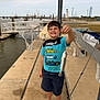 child, boy, smile, fish, dock, pier, water, harbor, boats, lighthouse, fishing_rod, catch, t_shirt, shorts, shoes, concrete, railing, sky, outdoor, happy