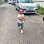 toddler, child, running, street, braided_hair, white_shoes, tshirt, tom_and_jerry, smiling, outdoor, cars, asphalt, sidewalk, greenery, urban, daylight, happy, playful, person, clothing