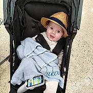 Lukas joined the competition — help win amazing prizes! baby, stroller, hat, blue_blanket, handheld_device, floor, happy, smiling, indoor, person, child, clothing, seatbelt, knitted_blanket, headwear, footwear, baby_clothes, technology, portrait, infant