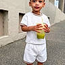 Chosyn is registered to the contest to win money with this photo: child, toddler, standing, portrait, outdoor, urban, building, concrete, fashion, white_outfit, shorts, sneakers, drink, cup, green_beverage, smiling, face, full_body, daylight, photography