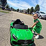 Chosyn joined the competition — help win amazing prizes! child, toddler, toy_car, green_clothing, outdoor, street, sunny, sky, clouds, vehicle, sneakers, sidewalk, residential_area, daytime, tree, car, road, play, kid, summer