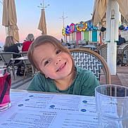 Dyana participe au concours pour gagner de l'argent avec cette photo : child, smiling, outdoor, cafe, table, drink, menu, glass, umbrella, evening, lights, happy, person, portrait, restaurant, seat, hair, face, clothing, necklace