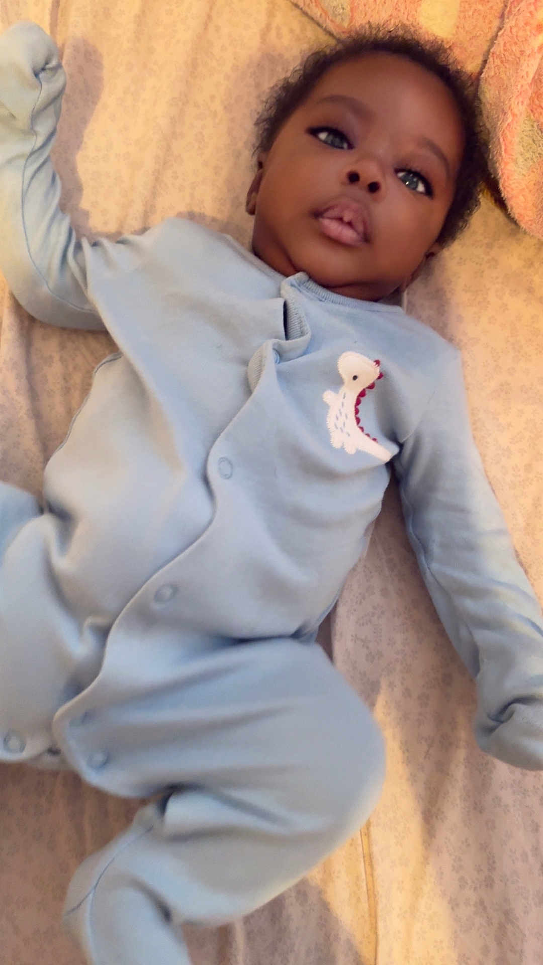 Pedro joined the competition — help win amazing prizes! baby, infant, onesie, blue_clothing, dinosaur_patch, bed, blanket, towel, lying_down, face, curious, indoors, soft_lighting, young_child, portrait, relaxed, child, sleepwear, person, cute