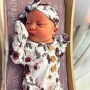 Evelyn joined the competition — help win amazing prizes! newborn, baby, infant, floral_outfit, headband, lying_down, bed, blanket, wooden_bed_frame, face, skin, clothing, button, purple, pattern, sleepwear, cute, portrait, indoors, person