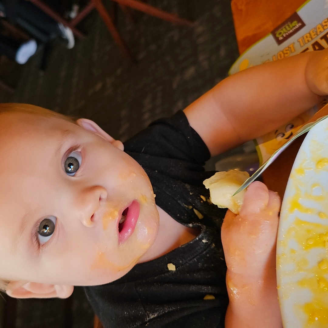 Finnigan joined the competition — help win amazing prizes! baby, black_shirt, child, curious_eyes, eating, face, food, fork, hand, high_chair, indoor, meal, messy_face, person, plate, restaurant, seat, table, toddler, wooden_chair