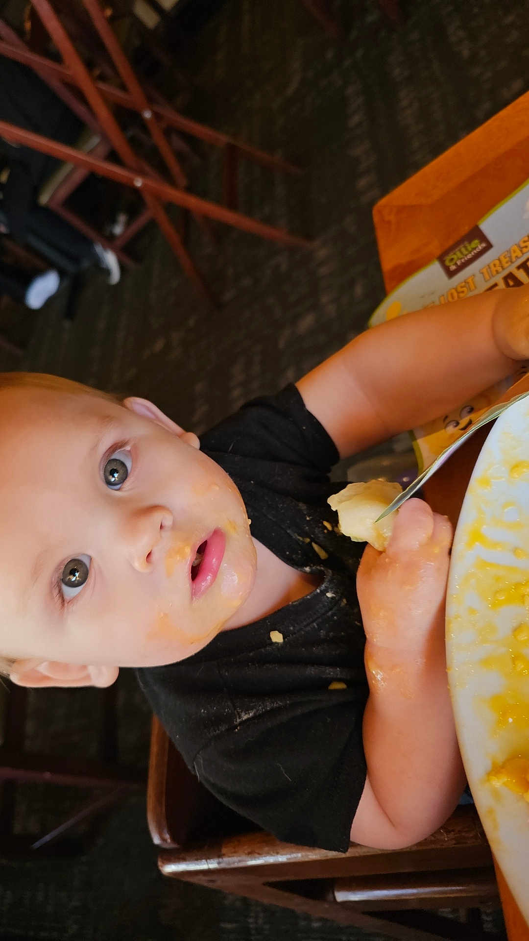 Finnigan joined the competition — help win amazing prizes! baby, black_shirt, child, curious_eyes, eating, face, food, fork, hand, high_chair, indoor, meal, messy_face, person, plate, restaurant, seat, table, toddler, wooden_chair