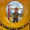 child, boy, playground, yellow_tunnel, smiling, outdoor, checkered_shirt, play_equipment, grass, trees, cloudy_sky, hands, face, happy, casual_clothing, daylight, park, fun, recreation, young_child