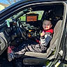 child, car, vehicle, driver_seat, smiling, open_door, computer_screen, sunlight, outdoor, tree, blue_sky, clothing, shoe, seat, dashboard, technology, person, happy, casual, daytime