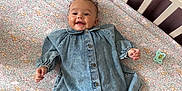 Jude is registered to the contest to win money with this photo: baby, denim_dress, crib, floral_sheet, toy, pacifier, smile, happy, infant, sleeping_area, fabric, child, furniture, soft_toy, pink_blanket, cute, indoors, person, face, legs
