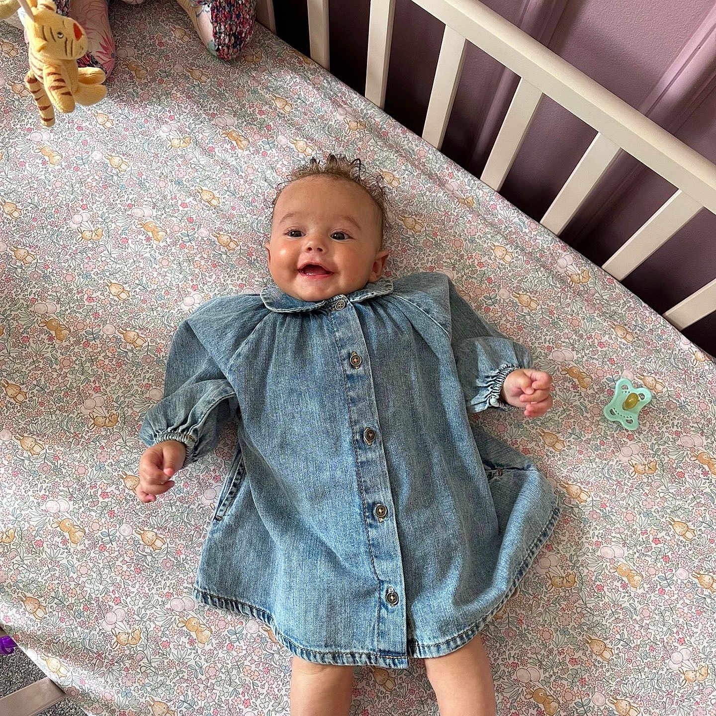 Jude is registered to the contest to win money with this photo: baby, child, crib, cute, denim_dress, fabric, face, floral_sheet, furniture, happy, indoors, infant, legs, pacifier, person, pink_blanket, sleeping_area, smile, soft_toy, toy
