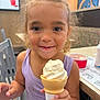 child, ice_cream, cone, smile, curly_hair, indoor, table, chair, food, vanilla, happy, person, casual_dining, face, hand, purple_clothing, dessert, young, cute, fun