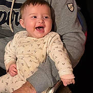 Lyah a rejoint le concours — aidez-le/la à gagner de superbes lots ! baby, child, adult, person, smiling, laughing, onesie, clothing, gray_hoodie, hand, chair, indoor, happy, cute, face, seated, parent, holding, joyful, expression