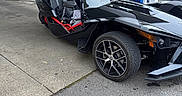 Star is registered to the contest to win money with this photo: dog, vehicle, three_wheeler, gas_station, sunglasses, black_dog, outdoor, pavement, wheel, fuel_pump, seat, road, tree, sky, building, sign, backpack, red_clothing, transportation, daytime