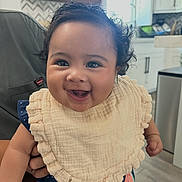 Angelene is registered to the contest to win money with this photo: baby, smiling, child, bib, curly_hair, indoor, kitchen, person, happy, cute, portrait, clothing, face, holding, infant, home, natural_light, furniture, cabinet, window