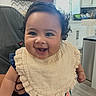 baby, smiling, child, bib, curly_hair, indoor, kitchen, person, happy, cute, portrait, clothing, face, holding, infant, home, natural_light, furniture, cabinet, window