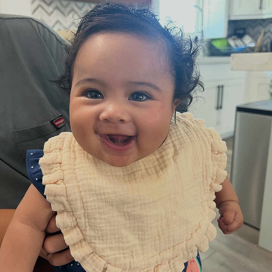 Angelene is registered to the contest to win money with this photo: baby, smiling, child, bib, curly_hair, indoor, kitchen, person, happy, cute, portrait, clothing, face, holding, infant, home, natural_light, furniture, cabinet, window