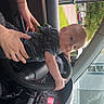 baby, child, car_interior, steering_wheel, hands, person, glasses, paper_towel, dashboard, seat, window, outside_view, greenery, daylight, clothing, infant, support, curious, portrait, vehicle