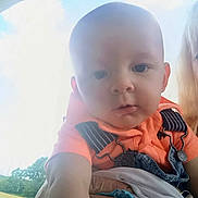 Julian is registered to the contest to win money with this photo: baby, child, infant, person, hand, clothing, orange_shirt, denim_overalls, outdoor, sky, clouds, greenery, portrait, closeup, curious, expression, holding, casual, daylight, face