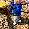 toddler, child, playground, wood_chips, clothing, blue_sweatshirt, gray_pants, sock, hands, face, sunlight, shadow, outdoor, person, play_equipment, curious, standing, daylight, casual, expression