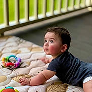 Grayson is registered to the contest to win money with this photo: baby, child, infant, mattress, toy, colorful_toy, patterned_bedding, dark_shirt, crawling, indoors, railing, green_background, curious_expression, wide_eyes, soft_light, skin, hand, face, head, floor