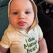 Elias is registered to the contest to win money with this photo: baby, child, hoodie, blue_eyes, indoor, cute, infant, shirt, text_on_clothing, portrait, person, sitting, young_child, face, expression, casual_clothing, hand, apparel, closeup, soft_lighting